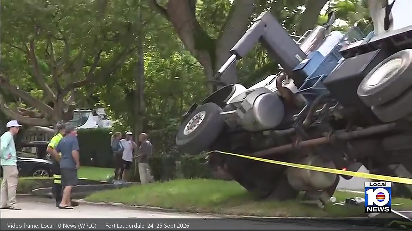 A boom truck lying on its side in the street after overturning, its outrigger beam, hydraulic tank and chassis exposed, yellow caution tape across the foreground