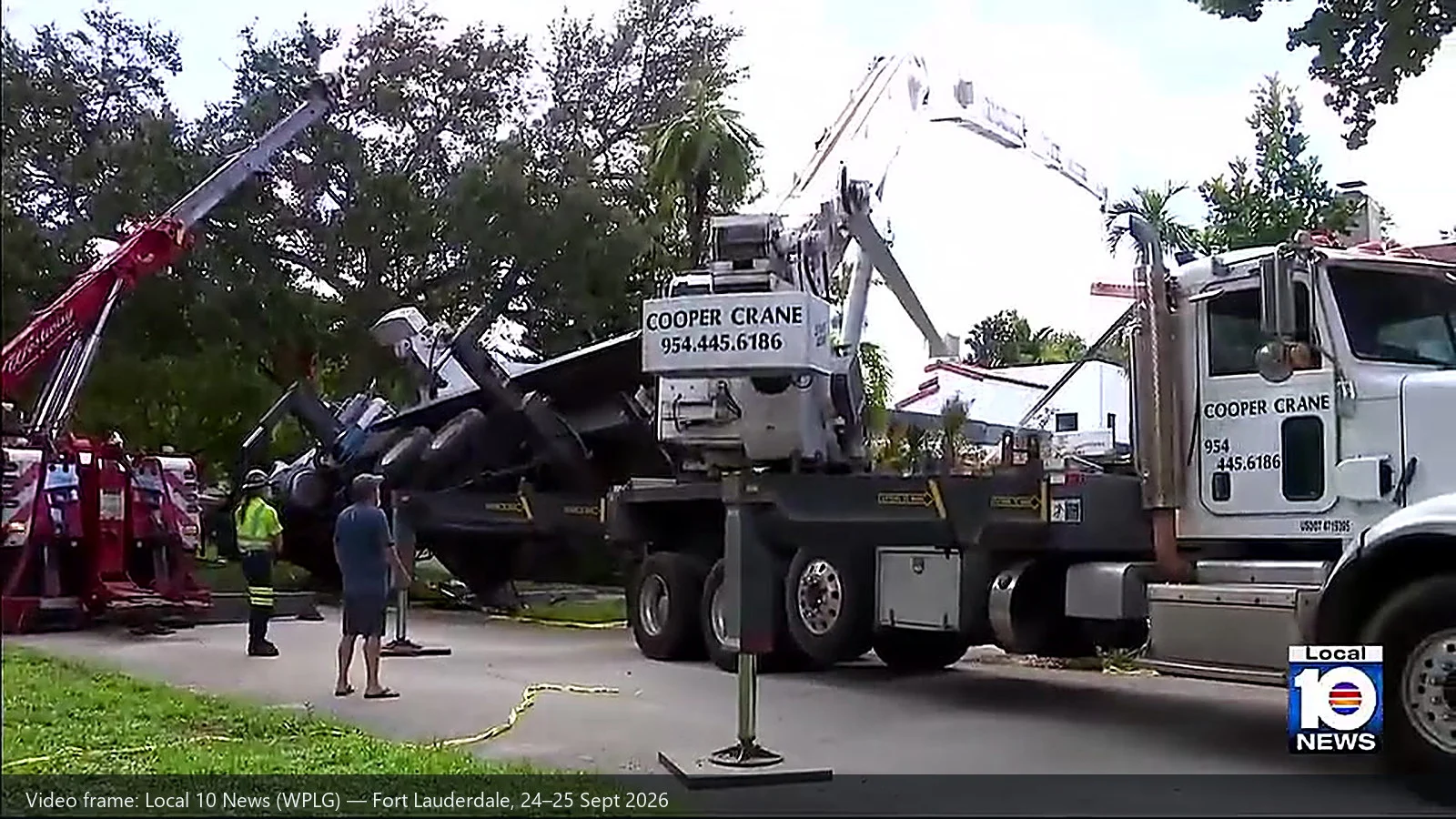 Recovery lift under way in a residential street: two crane trucks on either side and the overturned machine between them, with float pads and police tape on the ground
